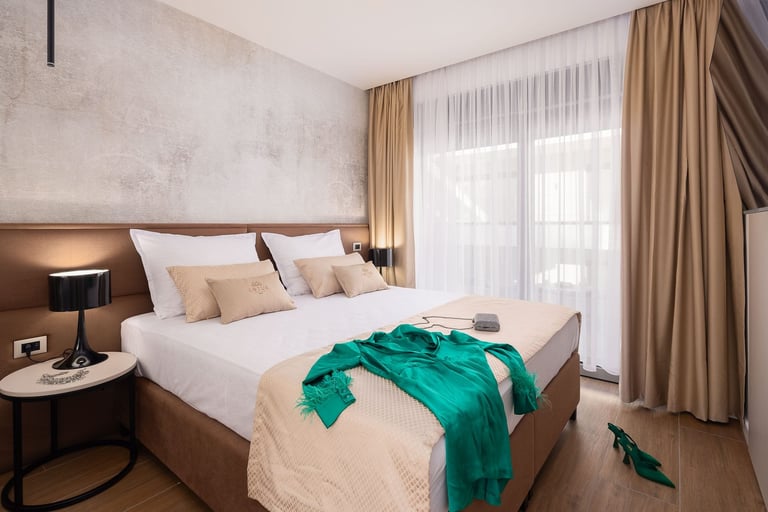 Modern bedroom with a large bed, green throw blanket, neutral bedding, wooden nightstands, floor-to-ceiling windows with beige curtains, and a textured accent wall