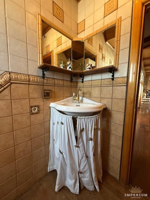 Bathroom corner with cream tile walls, corner sink with white skirt, mirrored cabinet above, and wooden door frame