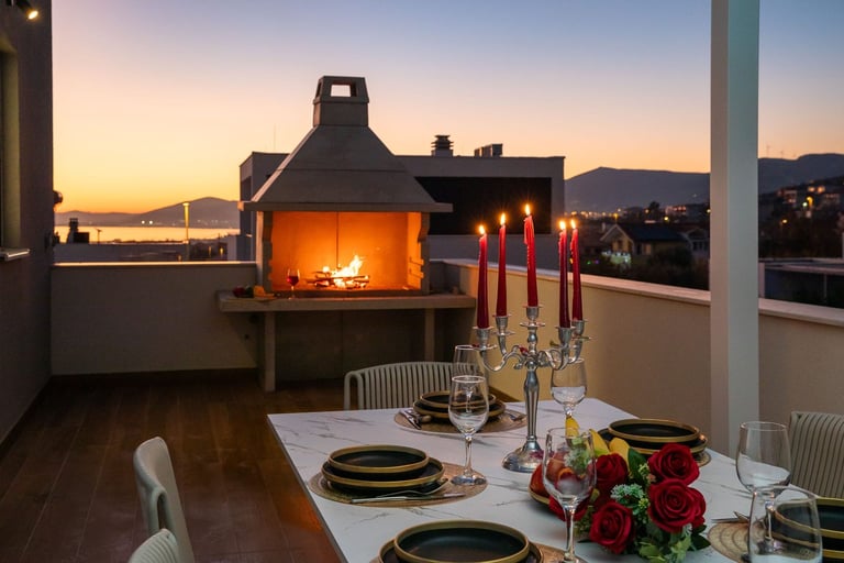 Elegant rooftop dinner setting with fireplace at sunset, decorated table with candles and red roses overlooking city lights