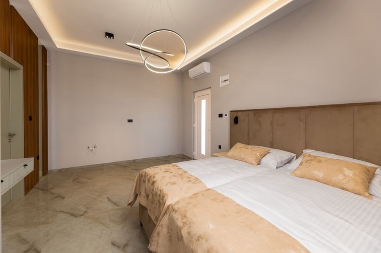 Modern bedroom with two twin beds, tan upholstered headboard, white bedding, orange accent pillows, and contemporary circular ceiling light fixture