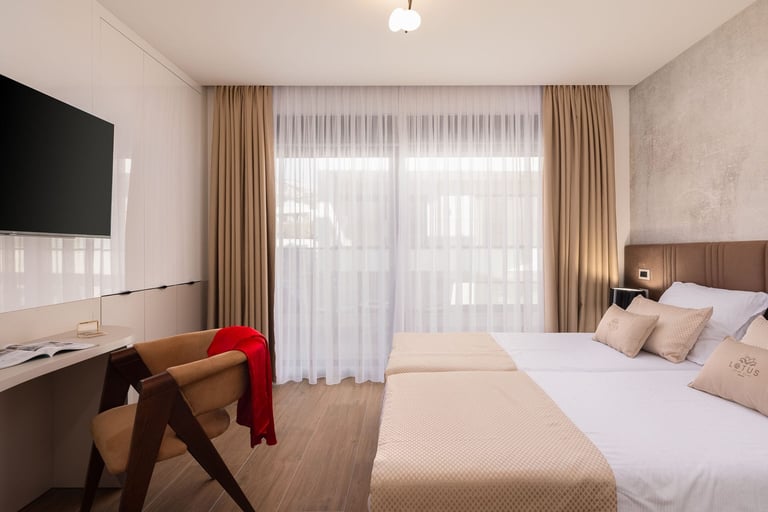 Modern hotel room with bed, desk with monitor, wooden furniture, and floor-to-ceiling windows with beige curtains
