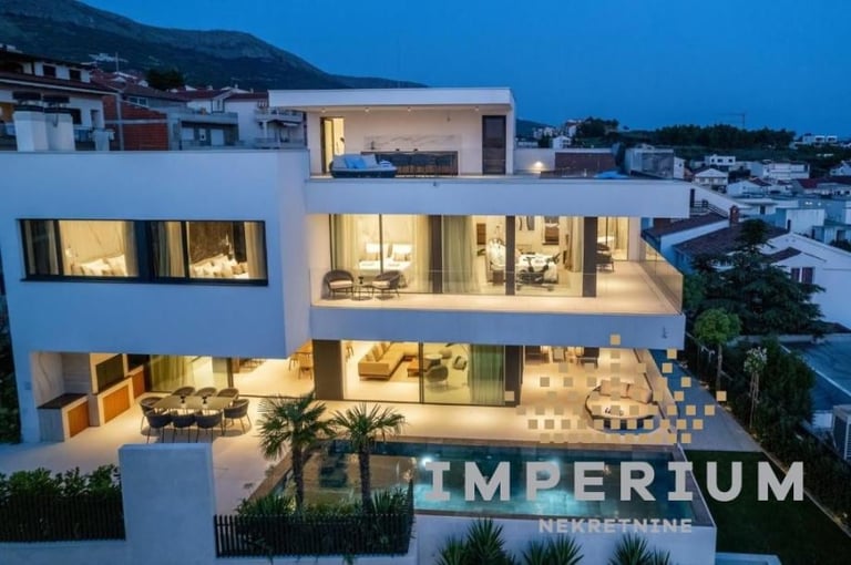 Modern white house with illuminated windows and Imperium signage at dusk in a Mediterranean coastal setting.