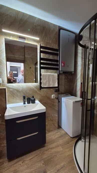 Modern bathroom with black vanity, white round sink, LED-lit mirror, beige tile walls, wooden floor, and glass shower enclosure