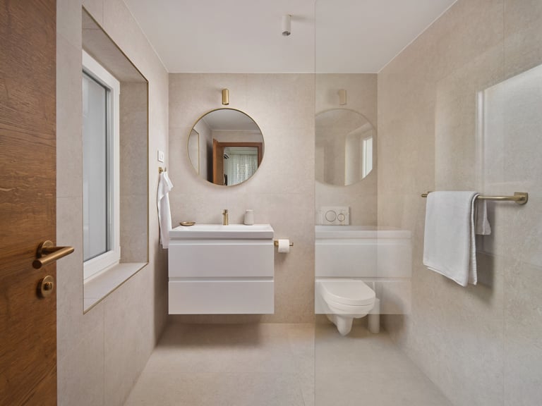 Modern bathroom interior with white vanity sink, round mirror, toilet, and shower enclosure with beige tile walls