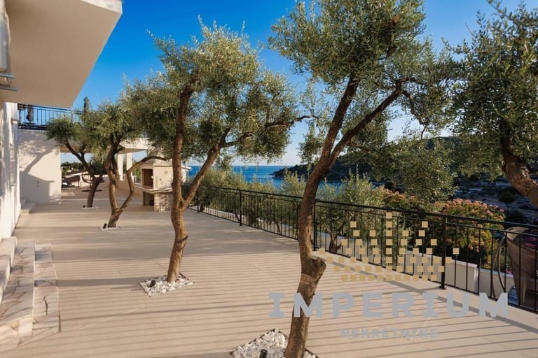 Coastal deck with olive trees overlooking blue Mediterranean sea, modern beige building, and metal railing on sunny day