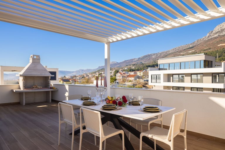 Modern rooftop terrace with dining table, chairs, pergola, mountain views, and built-in fireplace under clear blue sky