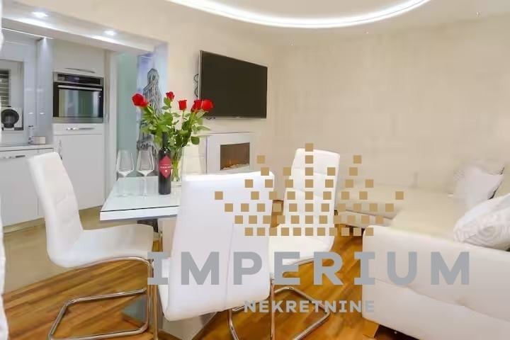 Modern studio apartment interior with open-plan living area, white furniture, kitchenette with appliances, mounted TV, and wooden flooring