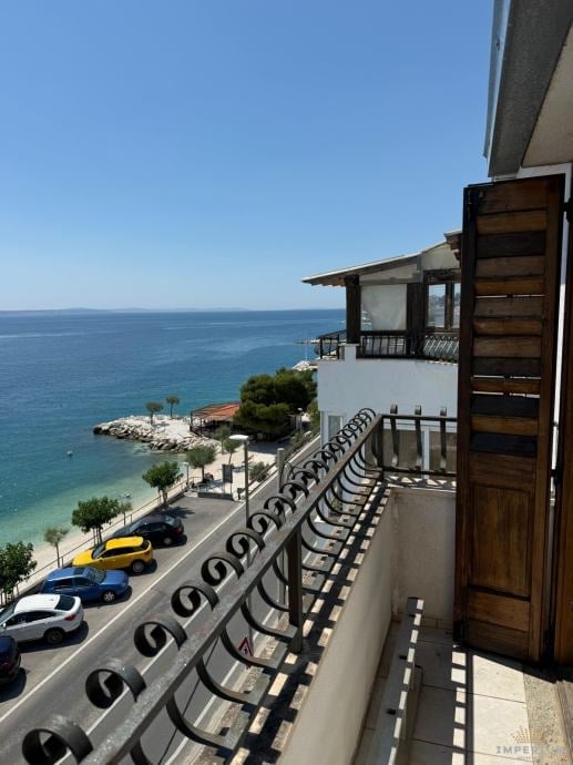 Coastal balcony view overlooking turquoise Mediterranean sea with lounge chairs, parked cars on beach road, and white waterfront buildings