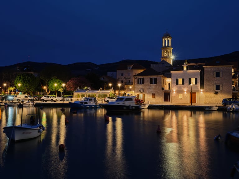 Nighttime harbor scene with illuminated waterfront buildings, boats reflected in calm blue water, and hills in background