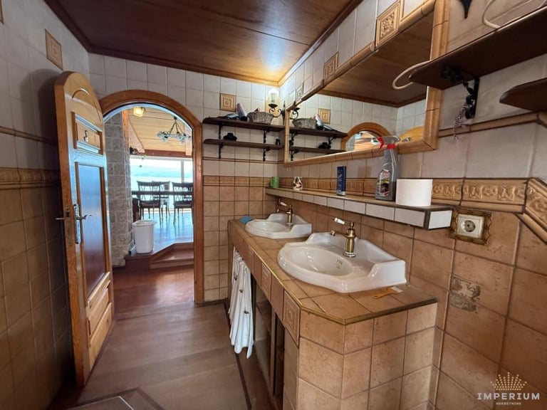 Spacious bathroom with dual sinks on wooden countertop, arched mirrors, brown tile walls, and wooden ceiling beams