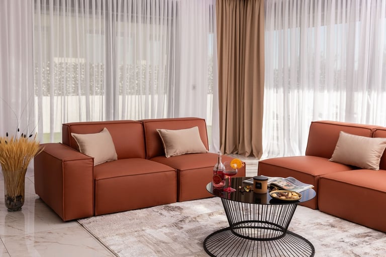 Modern living room with two burnt orange sofas, decorative pillows, and a black wire coffee table on a patterned rug, with sheer and brown curtains lining tall windows