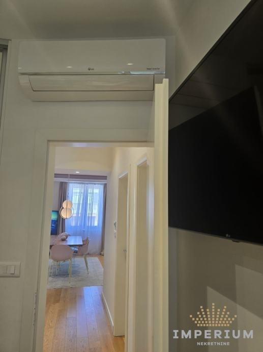 Modern hallway with white walls, mounted air conditioning unit, open doorway leading to living room with TV, and wooden flooring