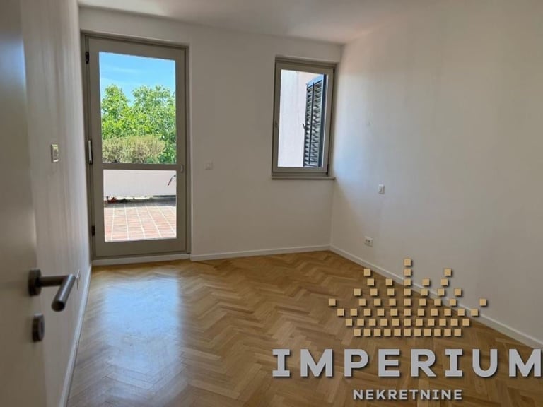Empty apartment room with herringbone wood flooring, glass door to balcony, window, and boxes stacked in corner