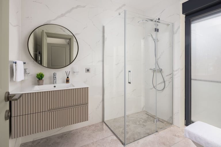 Modern bathroom with marble walls, round mirror above wooden vanity, glass shower enclosure, and chrome fixtures
