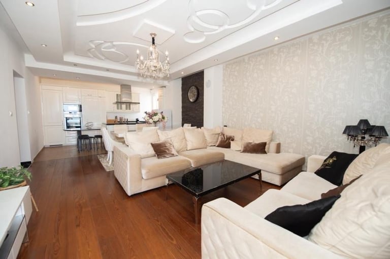 Modern open-plan living room with cream sectional sofa, black coffee table, wooden flooring, kitchen area, and decorative ceiling chandelier