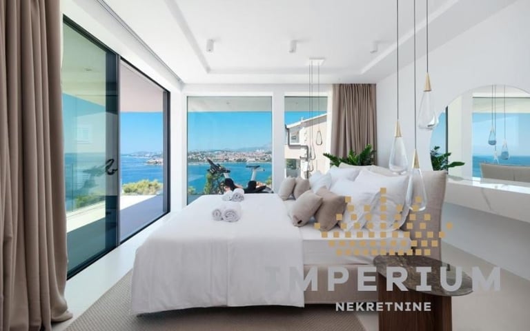 Modern bedroom with floor-to-ceiling windows overlooking a coastal city and blue water, white bed, and contemporary furnishings