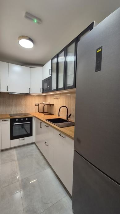 Modern kitchen with white lower cabinets, wooden countertop, black upper glass cabinets, built-in oven, and stainless steel refrigerator