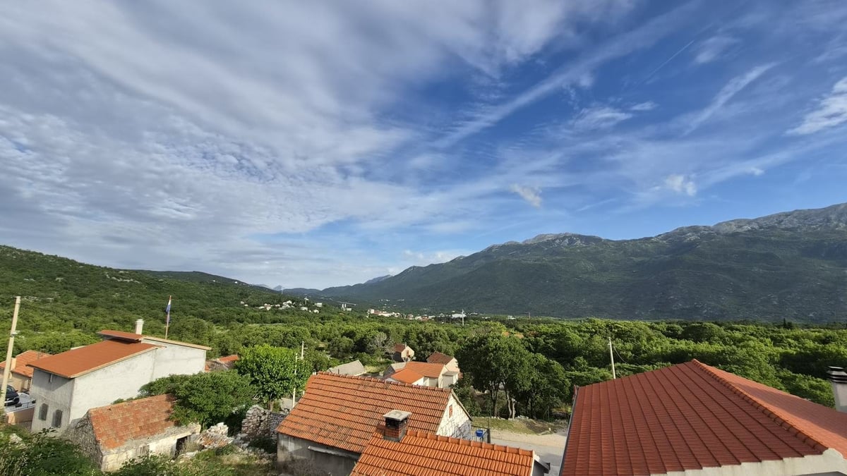 Aerial view of a Mediterranean village with red-tiled roofs, surrounded by green mountains and vineyards under blue sky