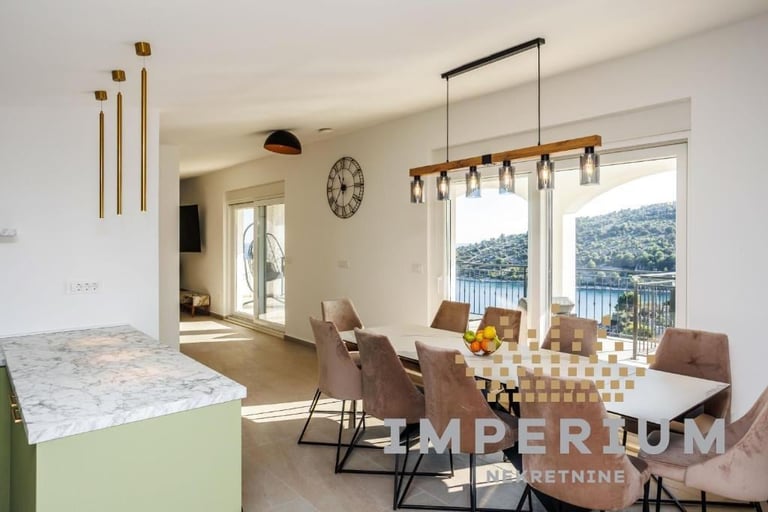Modern kitchen and dining room with lake view, featuring light green cabinetry, wooden table with brown chairs, and contemporary pendant lighting