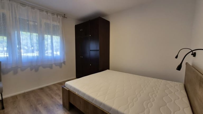 Bedroom with white quilted bed, dark wardrobe, window with sheer curtains, and wooden flooring