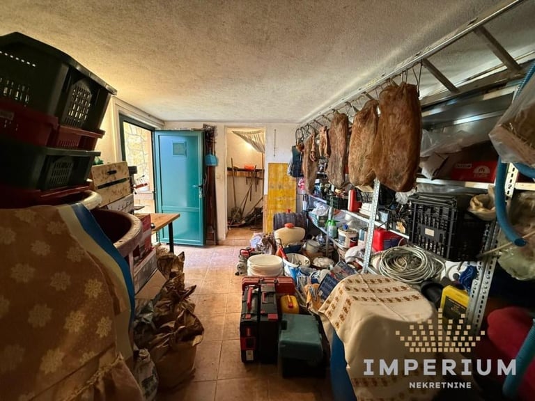 Cluttered storage room with shelving, hanging clothes, boxes, and various household items stacked throughout the space