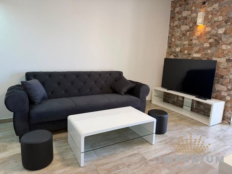 Modern living room with dark tufted sofa, white and glass coffee table, black ottoman, and flat-screen TV on brick accent wall