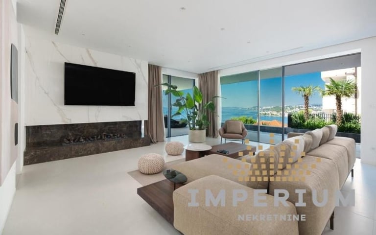 Luxury modern living room with beige sofa, wall-mounted TV, fireplace, and panoramic ocean views through floor-to-ceiling windows