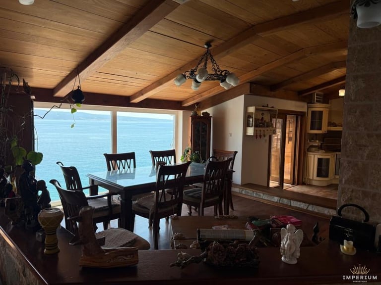 Spacious dining room with dark wood table, chairs, and ceiling beams, overlooking water through large windows