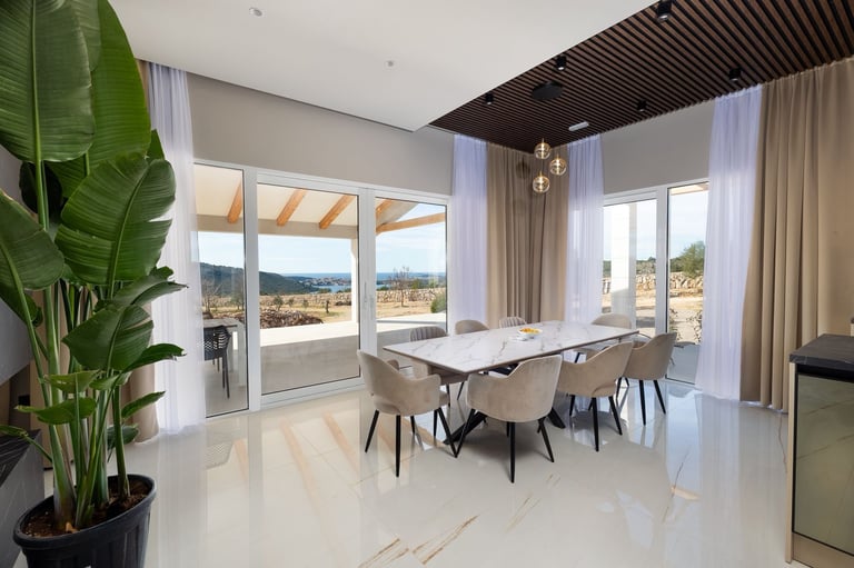 Modern dining room with floor-to-ceiling windows overlooking mountain landscape, white marble flooring, wooden table with beige chairs, and potted plants