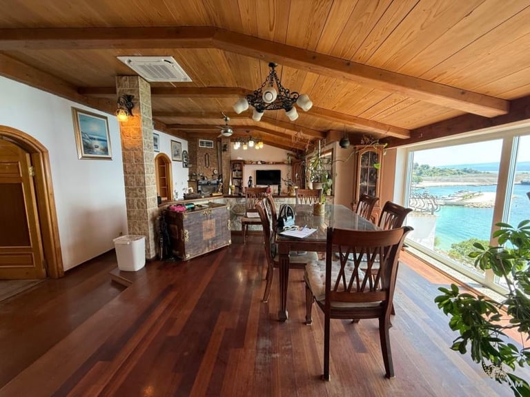 Spacious wooden interior with dining area, fireplace, and large windows overlooking turquoise water and mountains