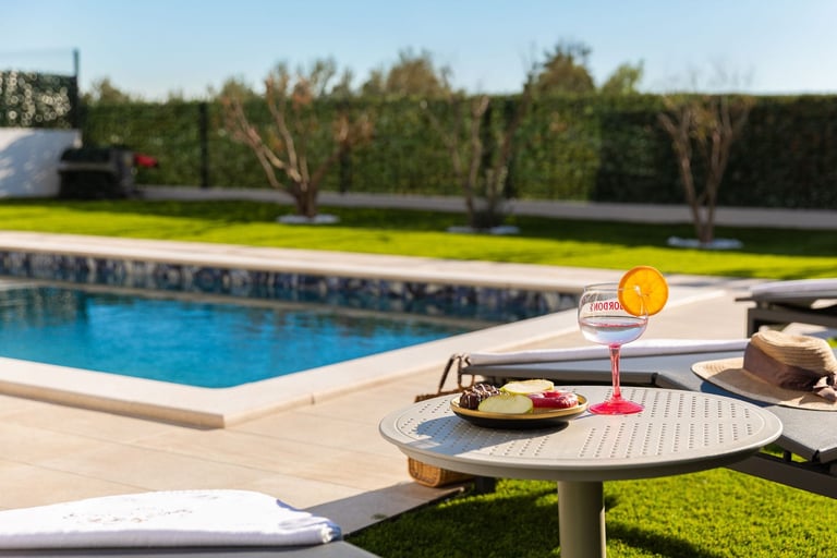 A poolside patio with a colorful cocktail and snacks on a table, overlooking a blue swimming pool and manicured lawn with landscaping