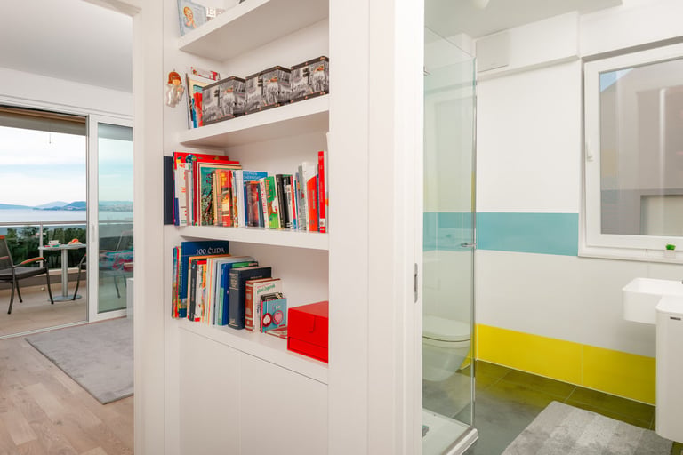 Modern apartment interior with white built-in shelving unit displaying books and red decorative boxes, bathroom visible with yellow and teal accents