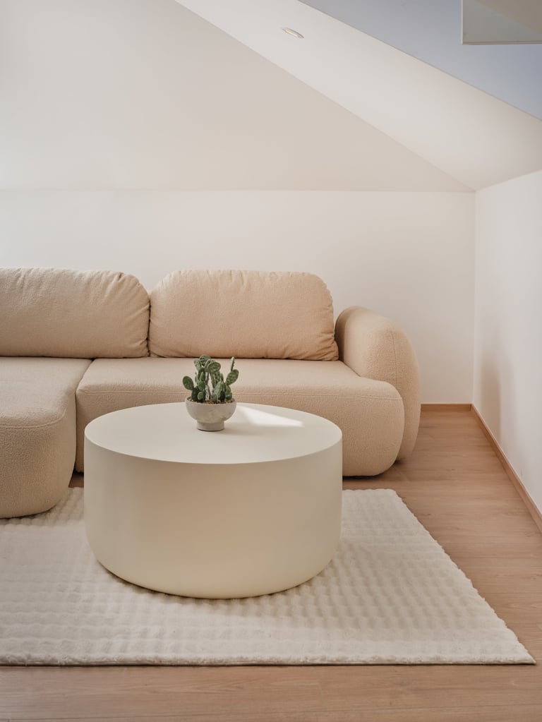Minimalist attic seating area with beige curved sofa, white cylindrical coffee table, small cactus, and cream rug on wooden floor