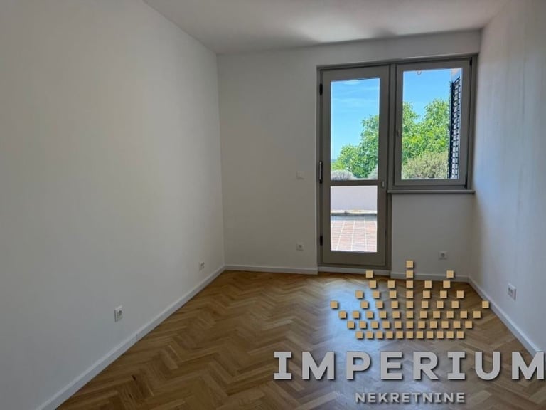 Empty apartment room with herringbone wood flooring, white walls, glass door and window, and cardboard boxes stacked in corner