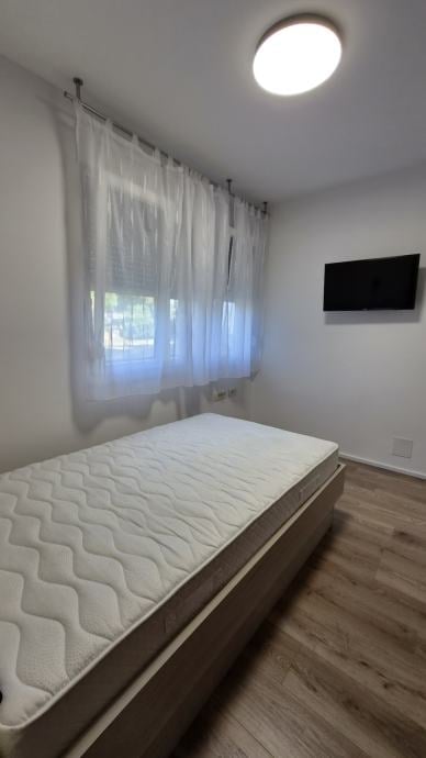 Minimalist bedroom with a platform bed, white sheer curtains on window, wall-mounted TV, round ceiling light, and wooden flooring.
