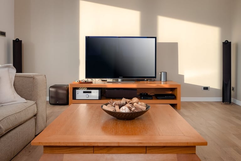 Modern living room with wooden furniture, flat-screen TV on stand, beige sofa, tall black speakers, and wooden table with bowl of nuts