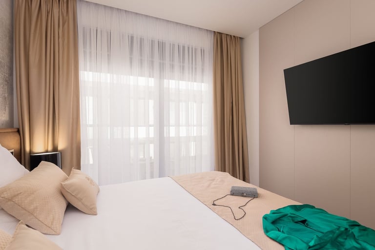 Modern hotel bedroom with white bed, beige pillows, wall-mounted TV, sheer and tan curtains, and a turquoise accent on the bed