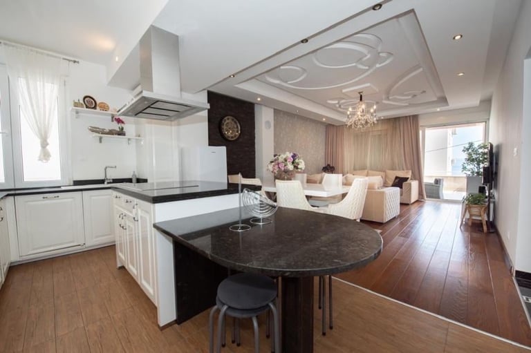 Modern open-plan kitchen and living room with white cabinetry, black granite island, hardwood floors, and contemporary ceiling design.