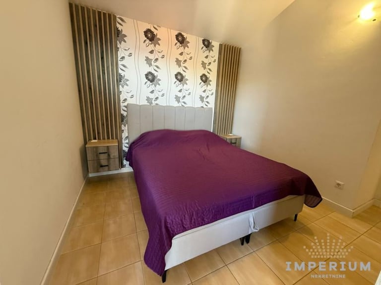 Modern bedroom with purple bed, white headboard with botanical artwork, wooden flooring, and beige walls with curtains