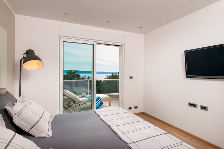 Modern bedroom with bed, sea view through glass doors, wall-mounted TV, and pendant lamp