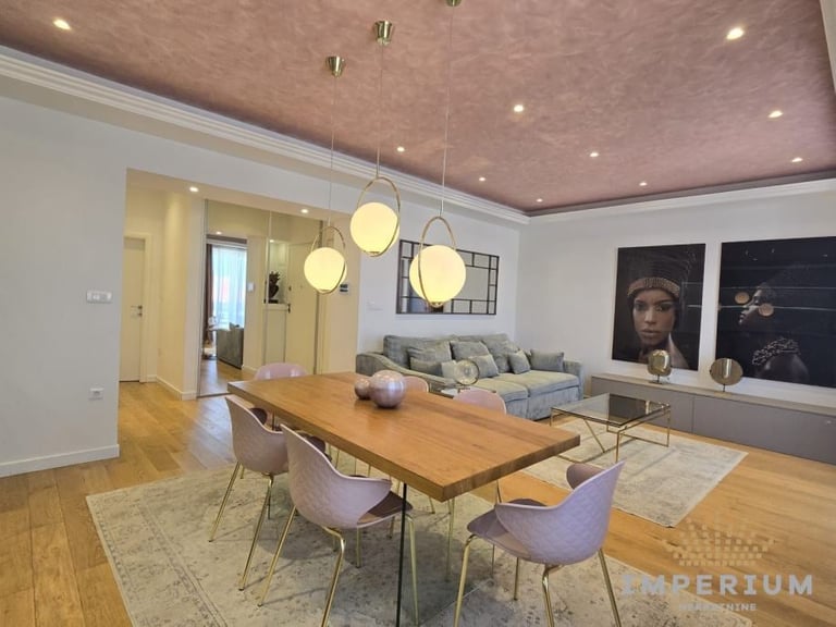 Modern dining room with wooden table, purple chairs, pendant lights, and grey sofa in background with decorative wall art.