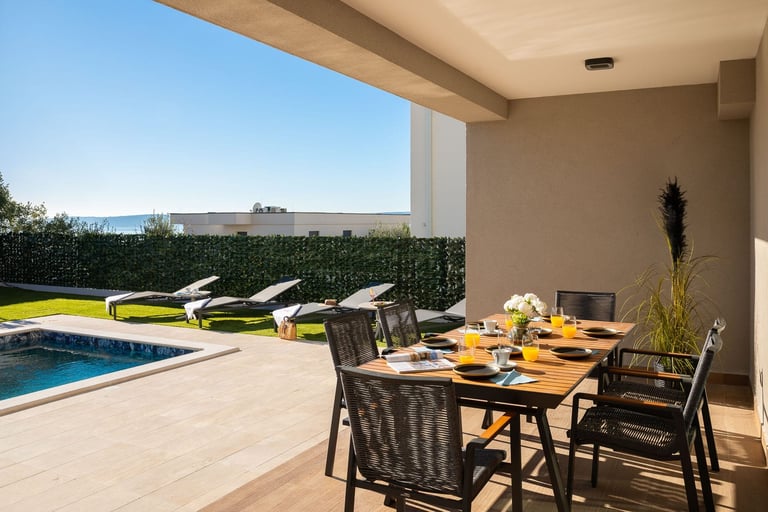 Modern patio with dining table, pool, loungers, and ocean view under a concrete pergola