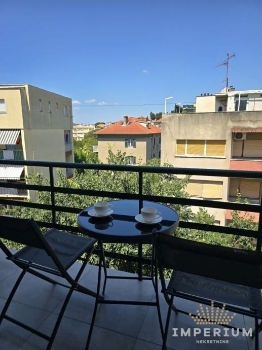 Balcony view overlooking a European urban neighborhood with apartment buildings, a small blue table with chairs, and clear blue sky