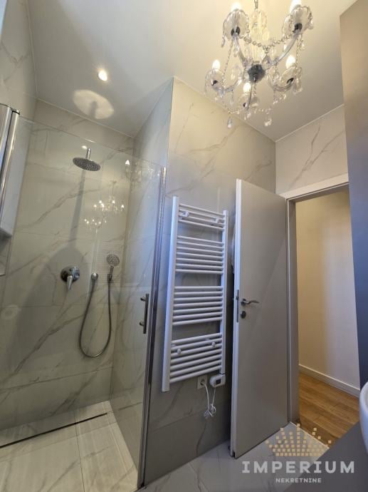 Modern luxury bathroom with marble walls, glass shower enclosure, heated towel rack, crystal chandelier, and marble flooring.
