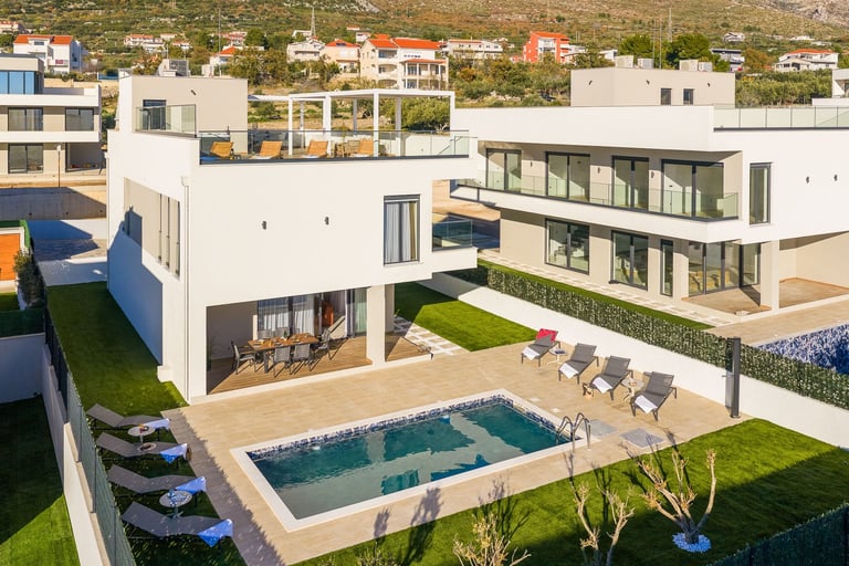 Modern white multi-level house with swimming pool, lounge chairs, green lawn, and surrounding residential neighborhood on hillside