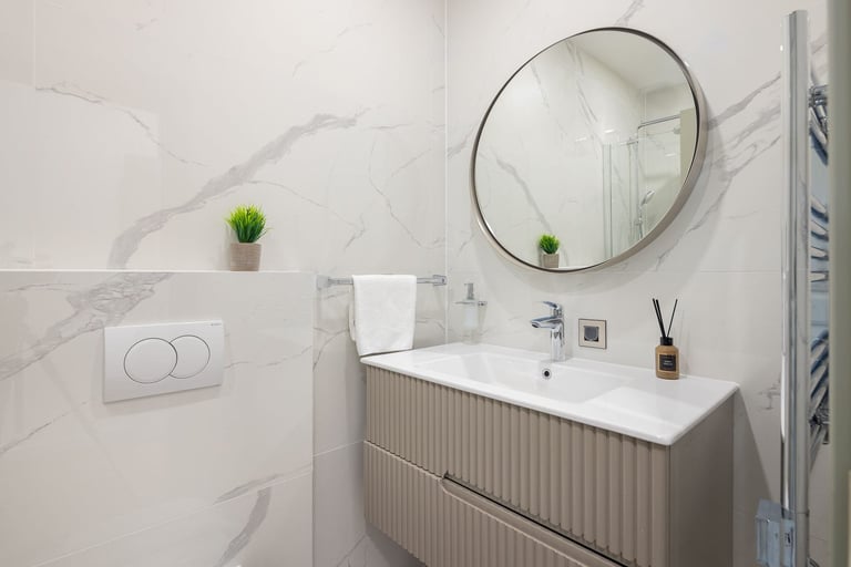 Modern bathroom with marble-effect tiles, round mirror, white sink vanity, and small potted plants on shelving