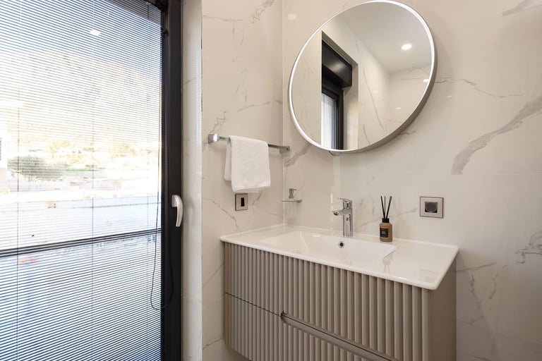 Modern bathroom with round illuminated mirror, white marble walls, ridged vanity cabinet, and window with blinds