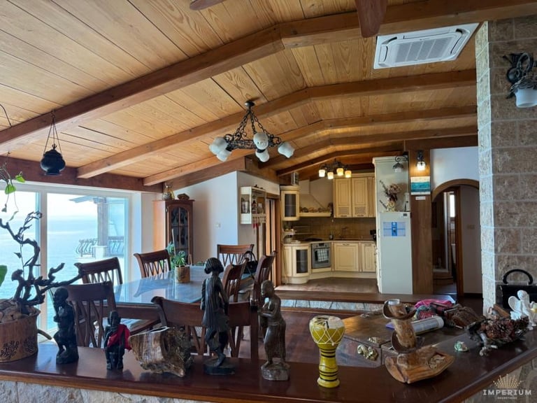 Rustic cabin interior with exposed wooden beams, dining area with dark chairs, kitchen in background, and stone fireplace on right wall