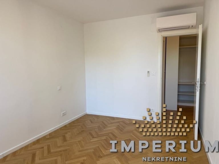 Empty apartment room with herringbone wood flooring, white walls, air conditioning unit, open closet door, and cardboard boxes