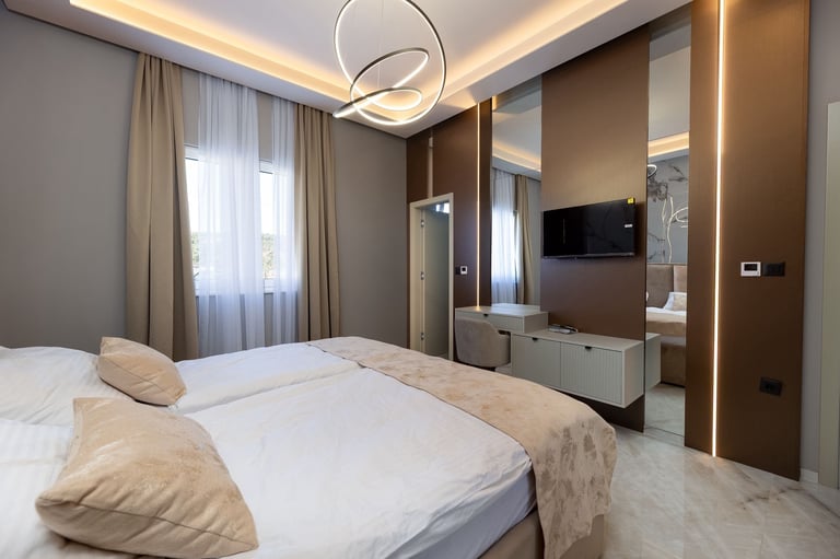 Modern hotel bedroom with white bed, warm brown walls, contemporary lighting, and mirrored wardrobe featuring integrated entertainment system